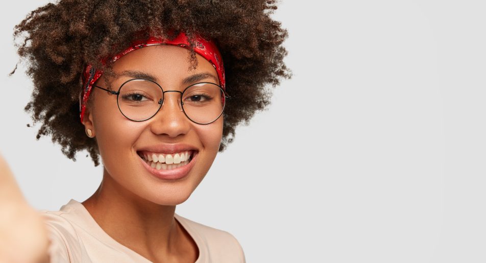 Close up shot glad feminine girl has toothy smile, Afro hairstyle, beautiful clean skin, wears transparent glasses, stretches hand as holds unrecognizable device, makes selfie over white background Close up shot glad feminine girl has toothy smile, Afro hairstyle, beautiful clean skin, wears transparent glasses, stretches hand as holds unrecognizable device, makes selfie over white background